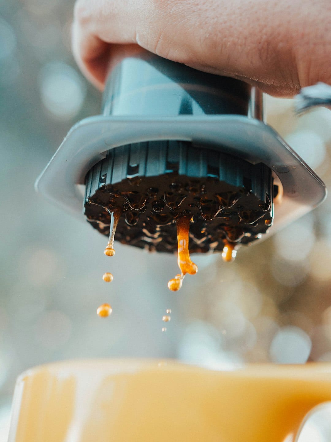 Close-up of an AeroPress filter cap as amber coffee drips into a mug, with a hand pressing the plunger and a bright outdoor background softened into bokeh.