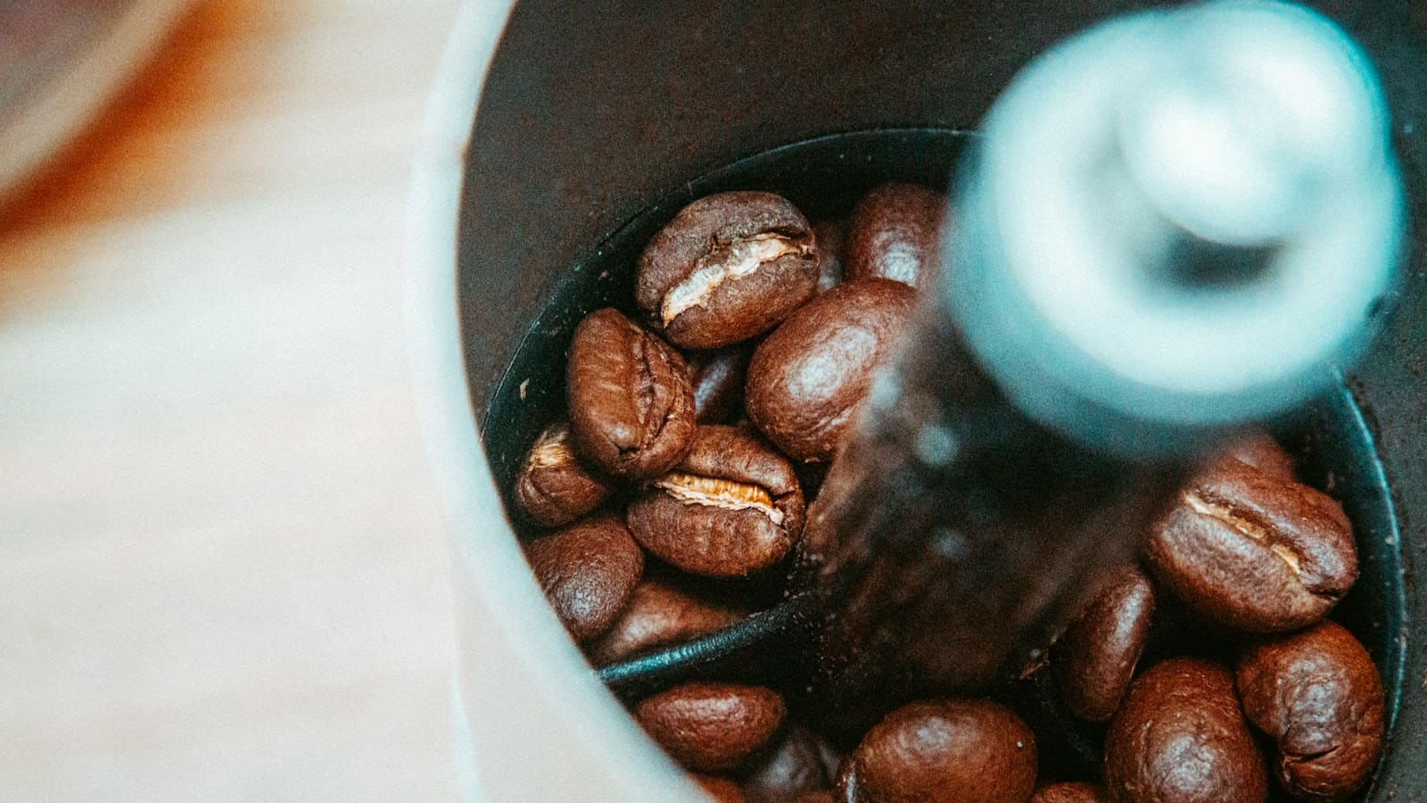 Top-down view into a coffee grinder hopper packed with roasted whole beans, the central axle soft in the foreground.