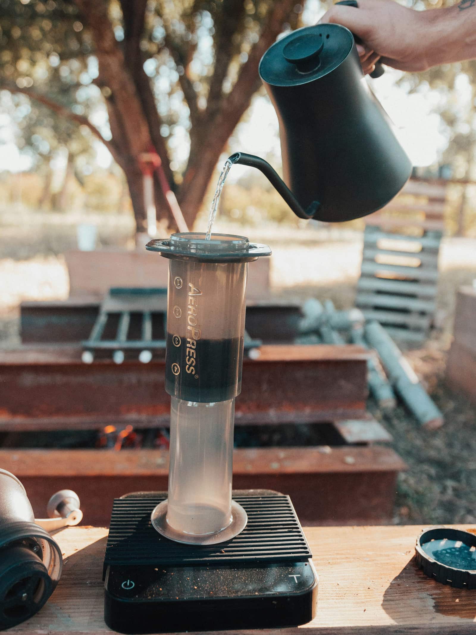 Outdoors, a hand pours water from a black gooseneck kettle into an inverted AeroPress standing on a coffee scale, with a hand grinder and fire pit nearby.