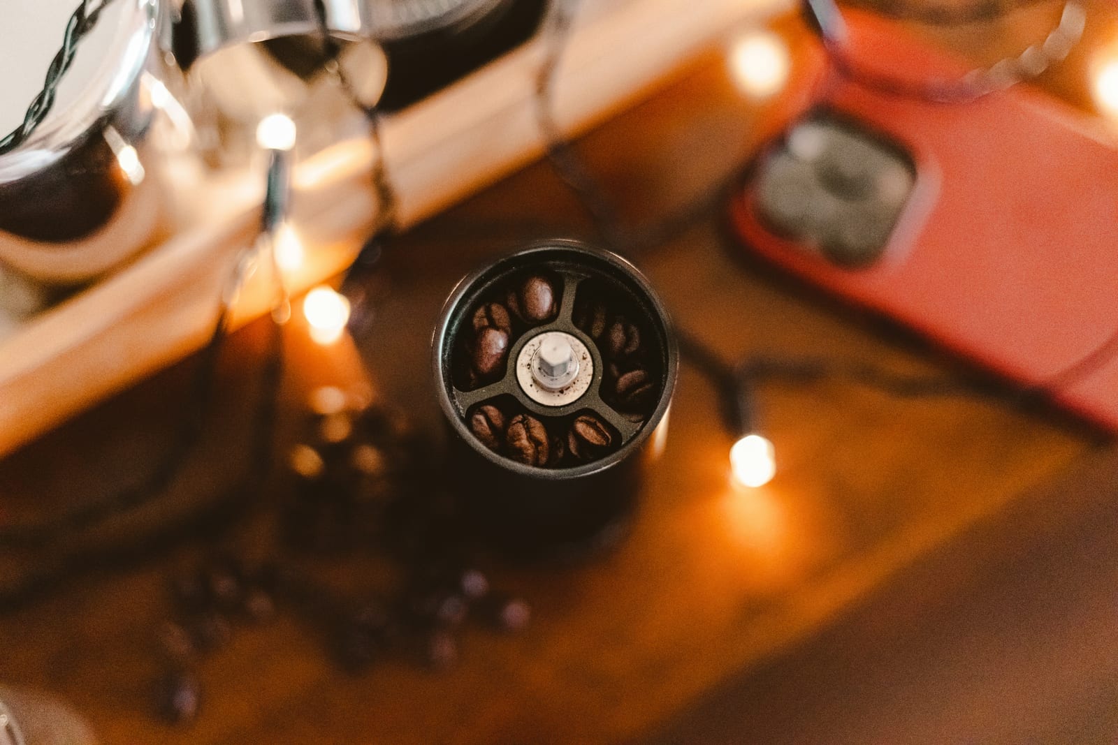 Looking down into a manual coffee grinder's hopper filled with roasted beans, standing on a wooden table with warm string lights around it.