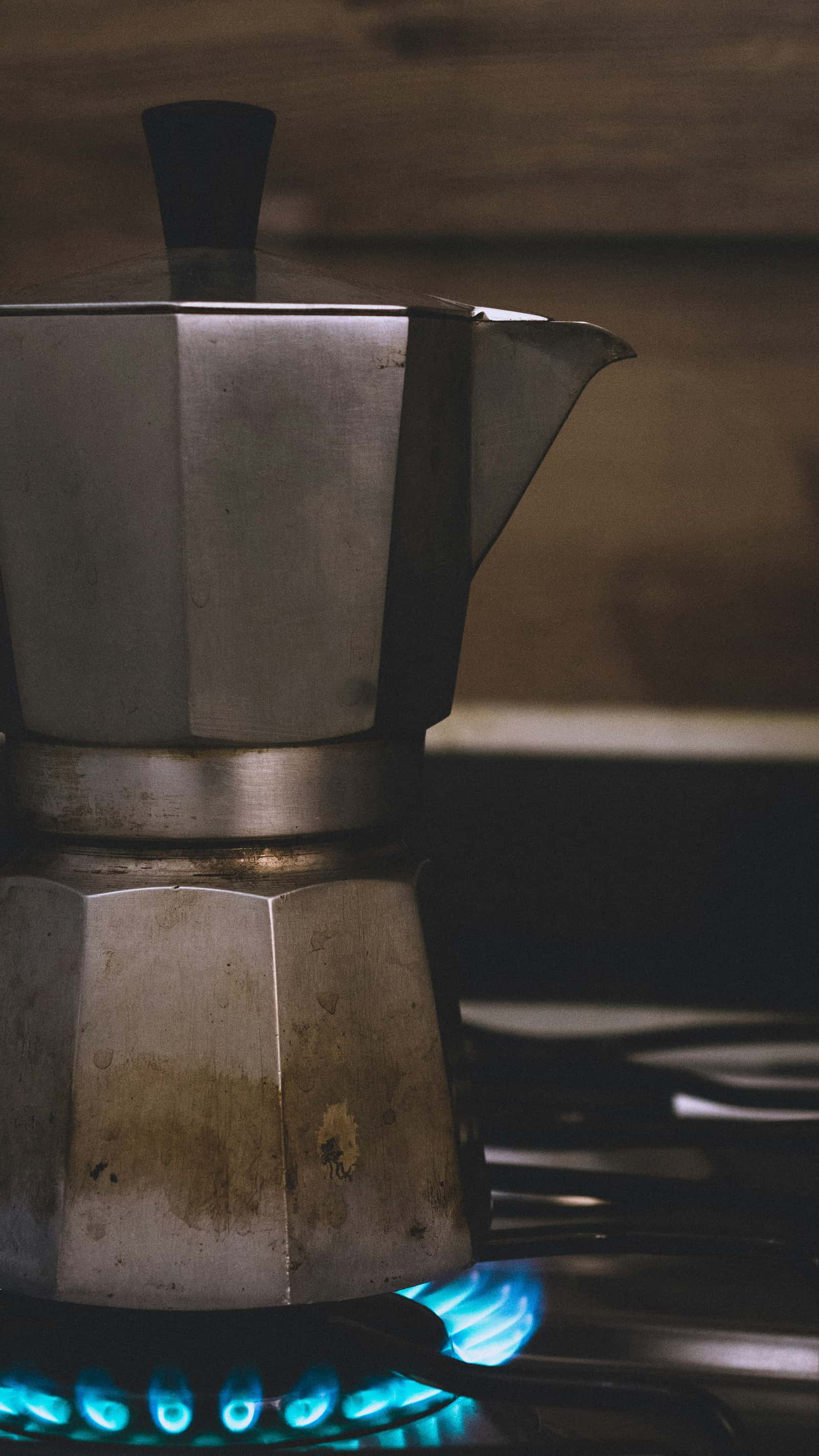 A well-used aluminum moka pot with a stained patina standing over a lit gas burner, blue flame visible under its base, in a dark kitchen.