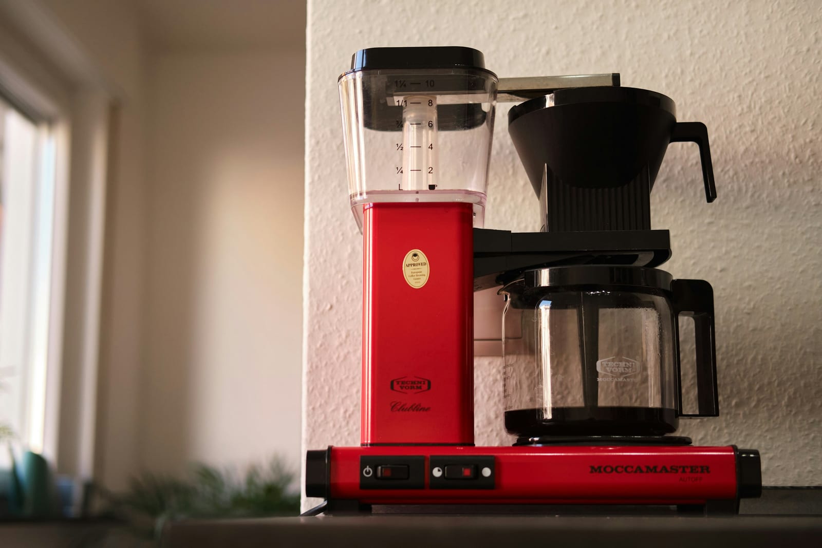 A red and black Technivorm Moccamaster automatic drip coffee machine on a counter, its gold certification seal visible on the water tower, with a blurred kitchen behind it.