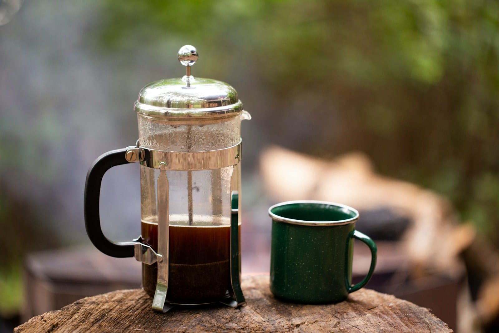 A glass French press full of brewed coffee with the plunger pushed down, next to a green enamel camp mug, both standing on a tree stump outdoors.