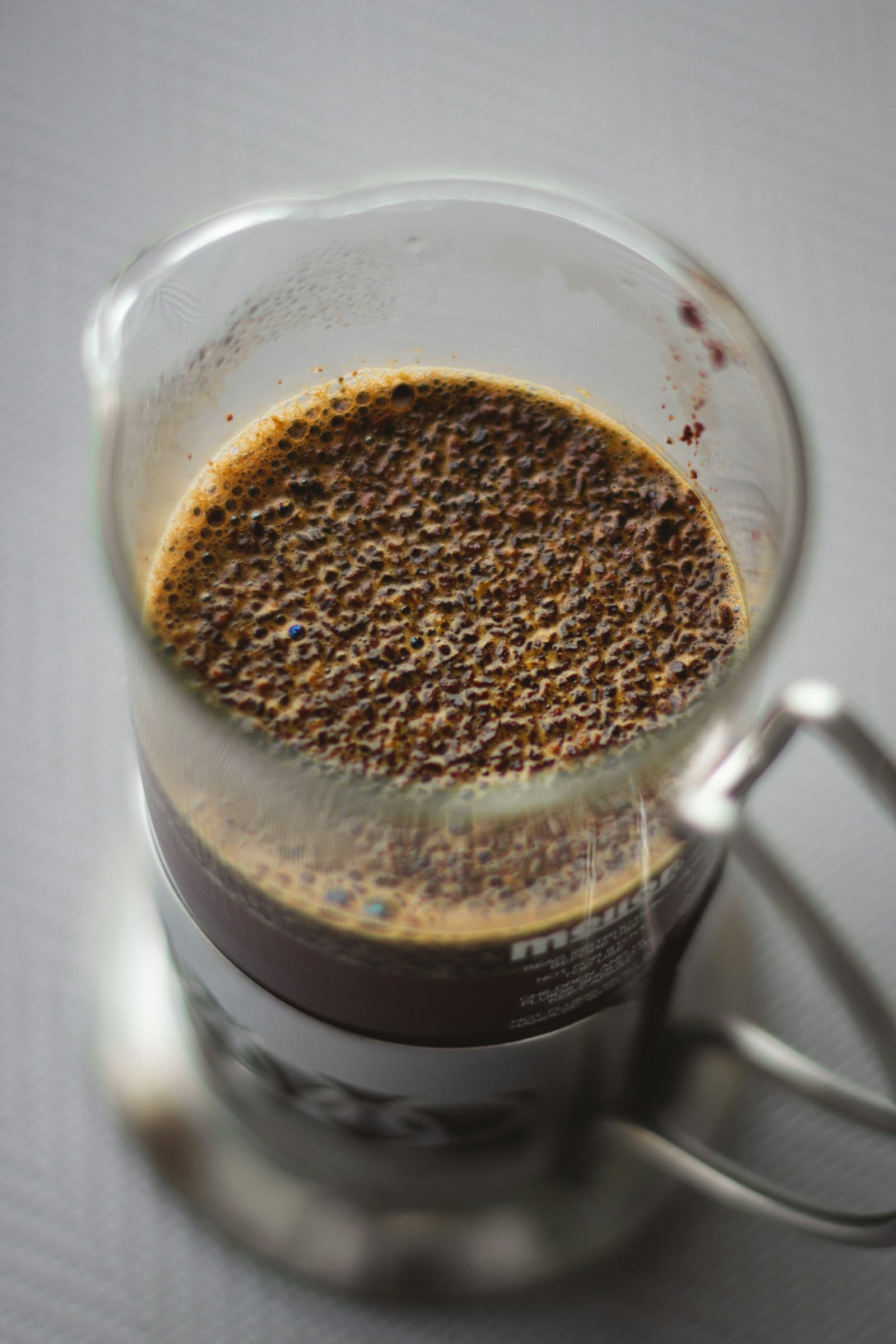Looking straight down into a French press mid-brew: a thick crust of wet coffee grounds floating on the surface, with oils and bubbles across the top.