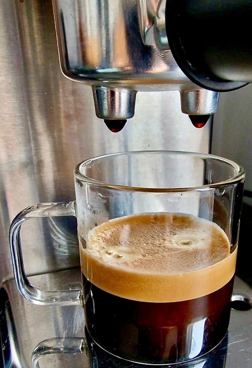 Primer plano de un shot de espresso a mitad de la extracción. Las dos bocas del portafiltro sobre una taza de vidrio, espresso oscuro con una crema dorada y espesa formándose encima.