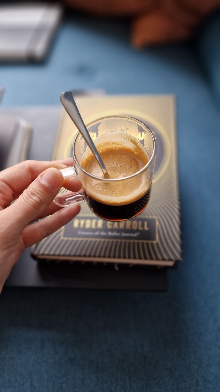 Una mano sosteniendo una tacita de vidrio de espresso con crema, apoyada sobre un ejemplar de The Bullet Journal Method, de Ryder Carroll, en un sofá azul.