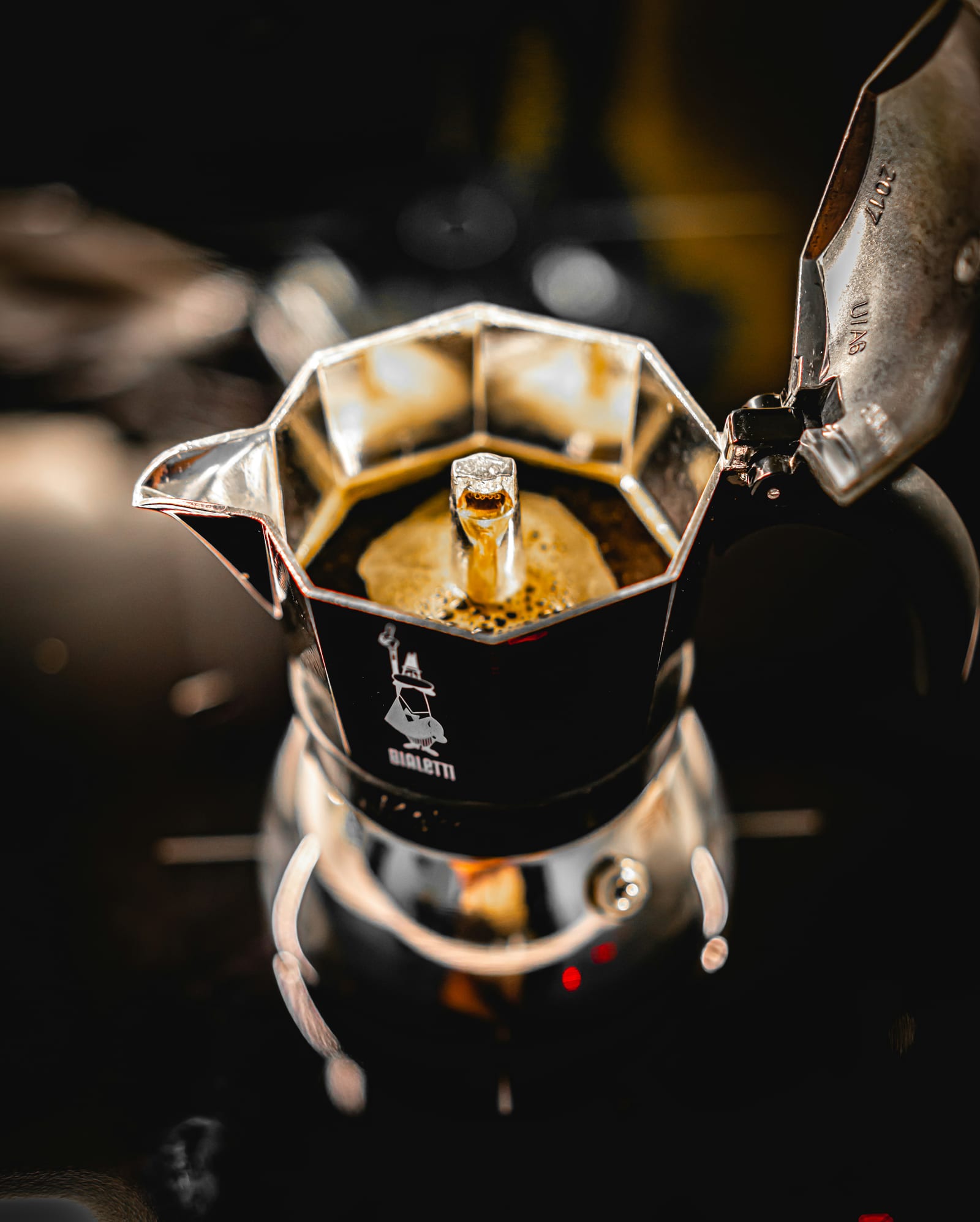 Una clásica cafetera moka Bialetti Moka Express con la tapa abierta, mostrando el café burbujeando hacia arriba por la chimenea central hasta la cámara superior, con el logo de Bialetti visible en el lateral.
