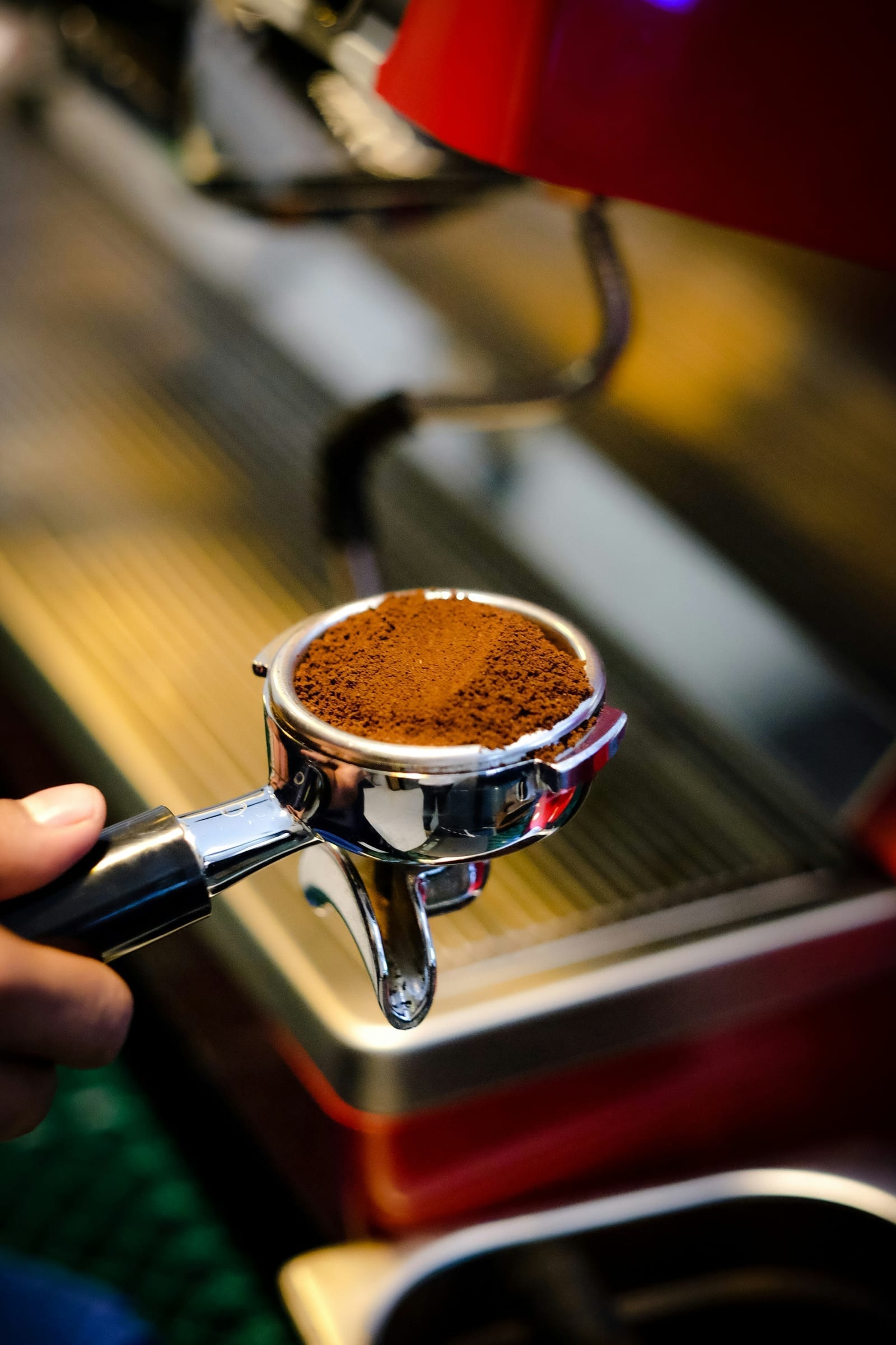 Una mano sosteniendo un portafiltro cromado de espresso lleno de café recién molido, delante de la bandeja de goteo de una máquina de espresso, con un molino rojo visible al fondo.