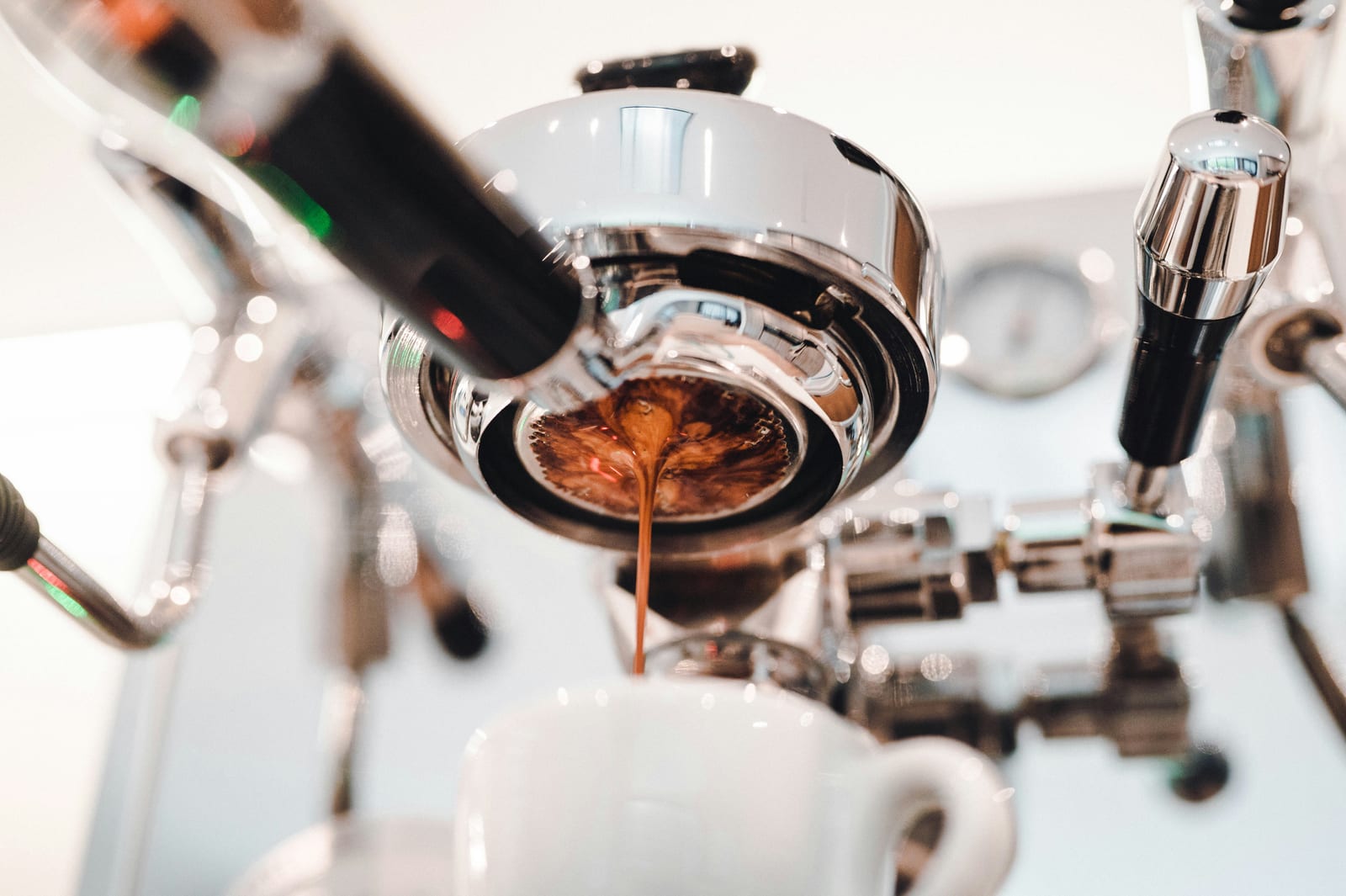 Un portafiltro sin fondo bajo el grupo cromado de una máquina de espresso sacando un chorro grueso y marrón de espresso a una taza blanca debajo, con un manómetro visible detrás.
