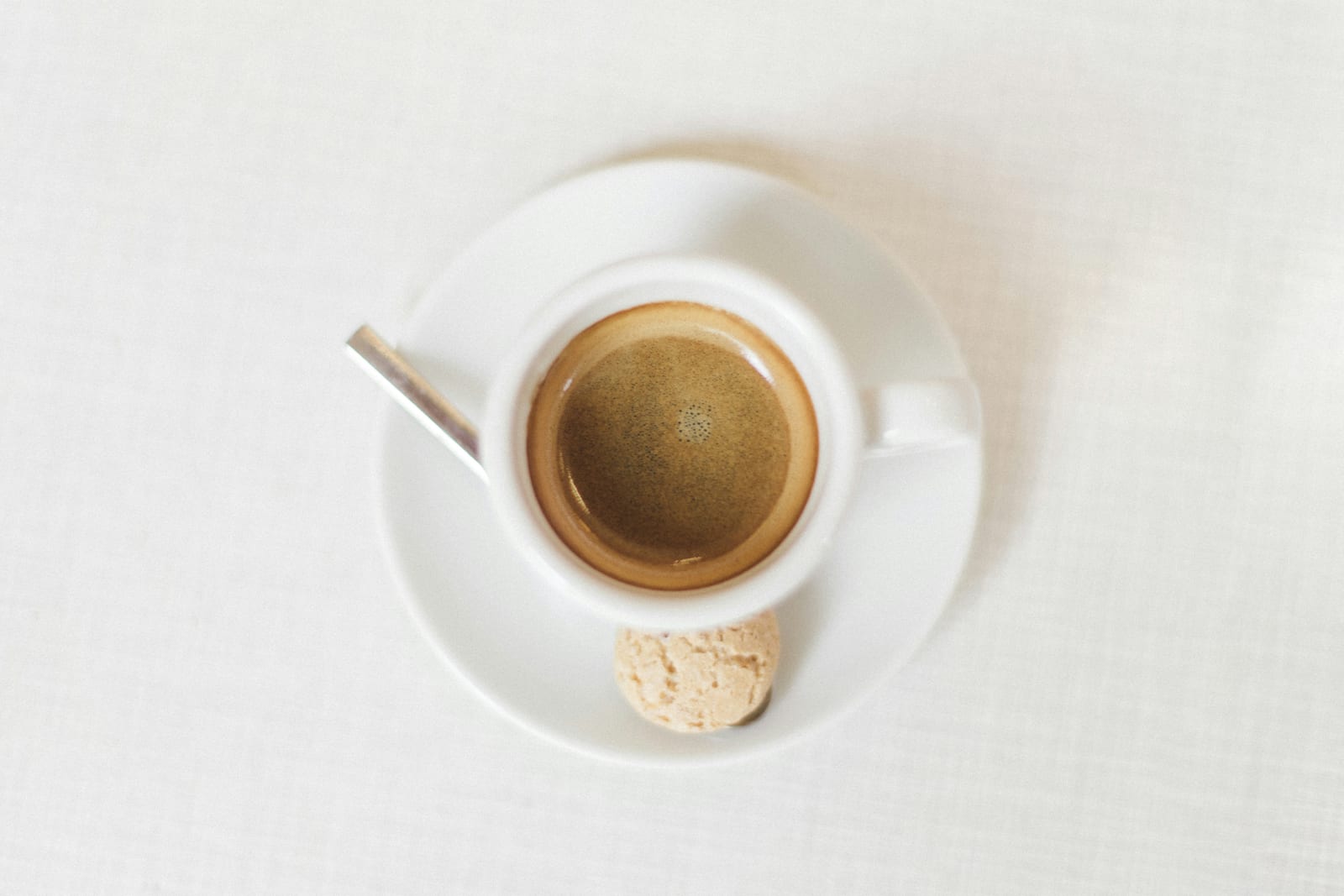 Overhead view of an espresso in a small white cup on a saucer, with a thick layer of golden crema, a silver spoon, and an amaretti cookie on the side.