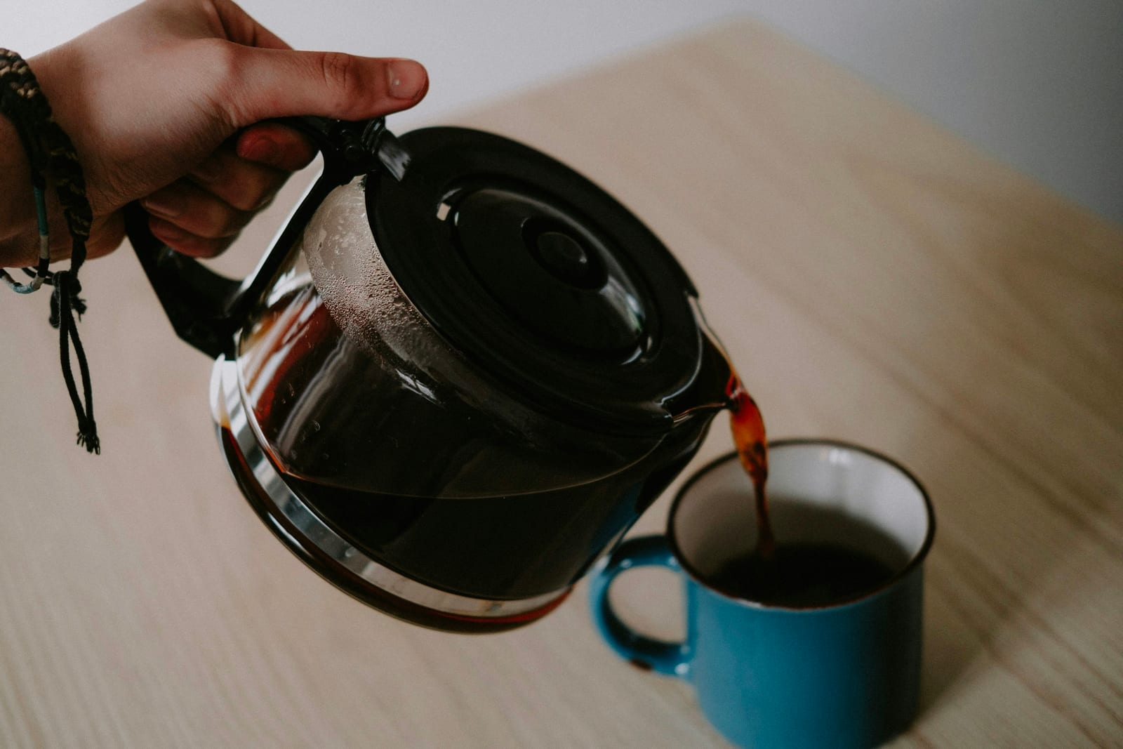 A hand tilts a full glass drip carafe with a black lid, pouring brewed coffee into a blue enamel mug on a pale wooden table.