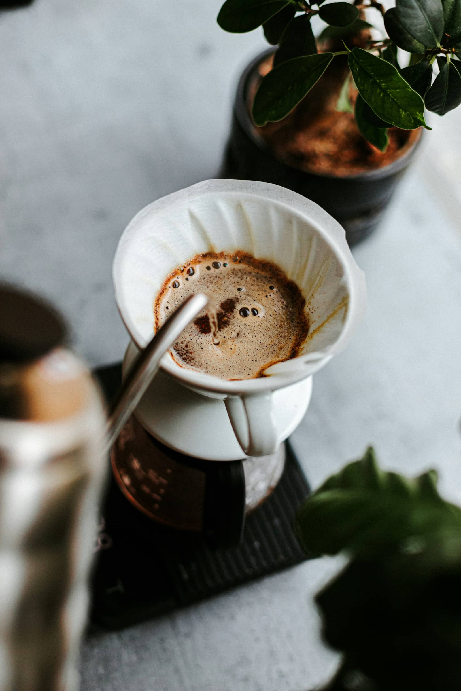 Looking straight down into a pour-over cone: wet coffee grounds foaming and doming inside a white paper filter, a steel kettle spout entering the frame, the dripper standing on a scale.