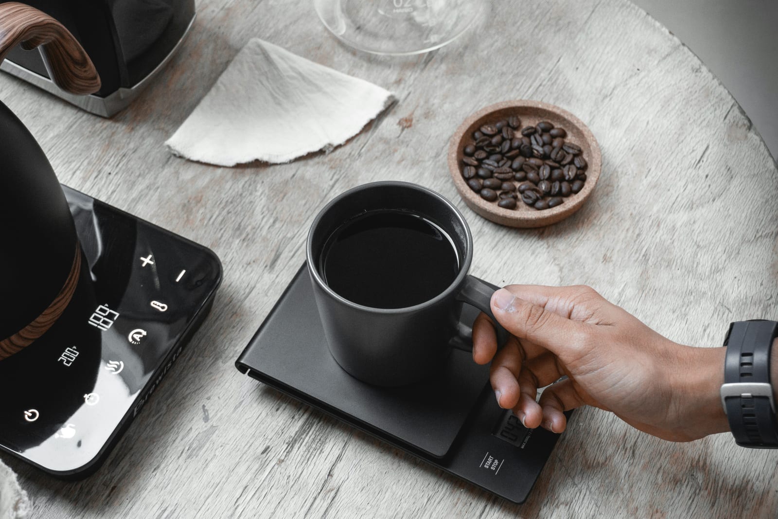 Overhead view of a brewing setup: a hand placing a black mug on a digital scale, an electric kettle showing its temperature display, a small wooden dish of coffee beans, and a paper filter resting nearby.