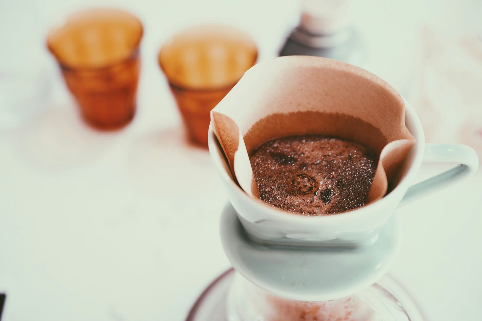 A paper-filtered pour-over dripper with the coffee bloom bubbling on top of the grounds, two amber glass cups waiting in the soft-focused background.