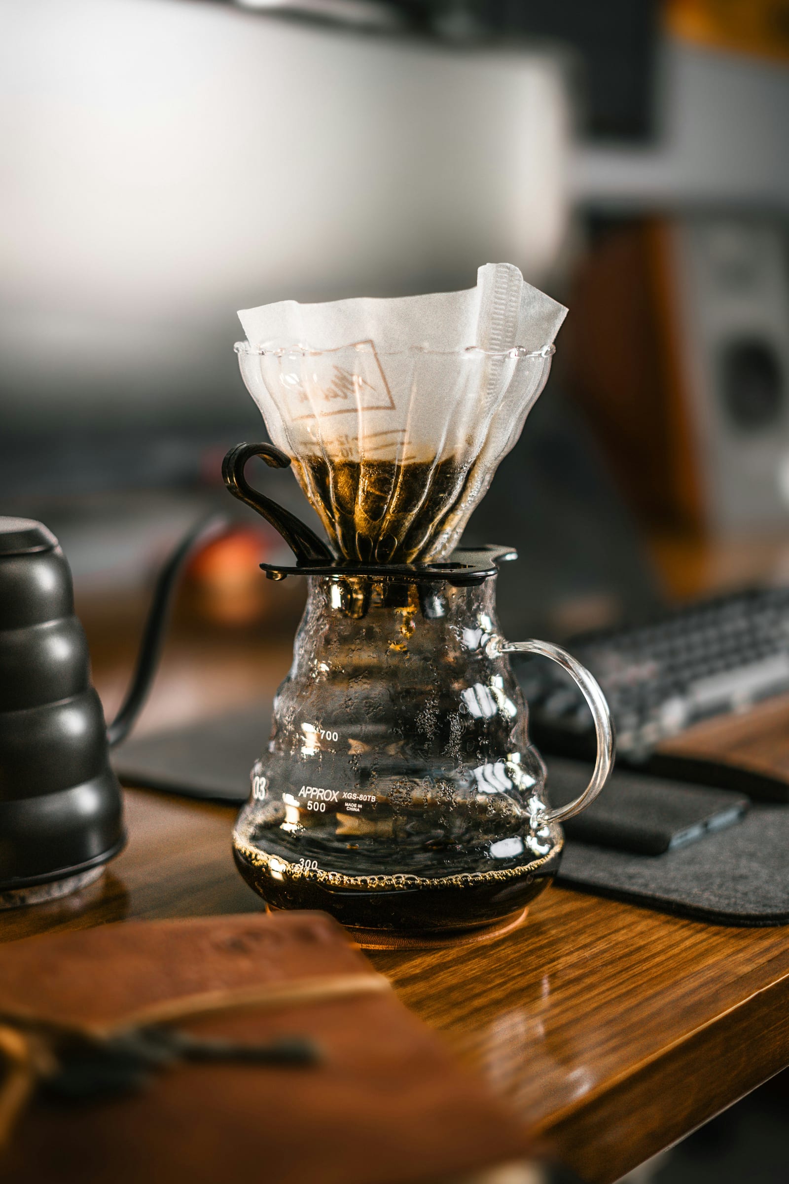 A V60 pour-over dripper with paper filter sits on a glass server on a wooden desk, with dark brewed coffee already collected below and a gooseneck kettle visible on the left.