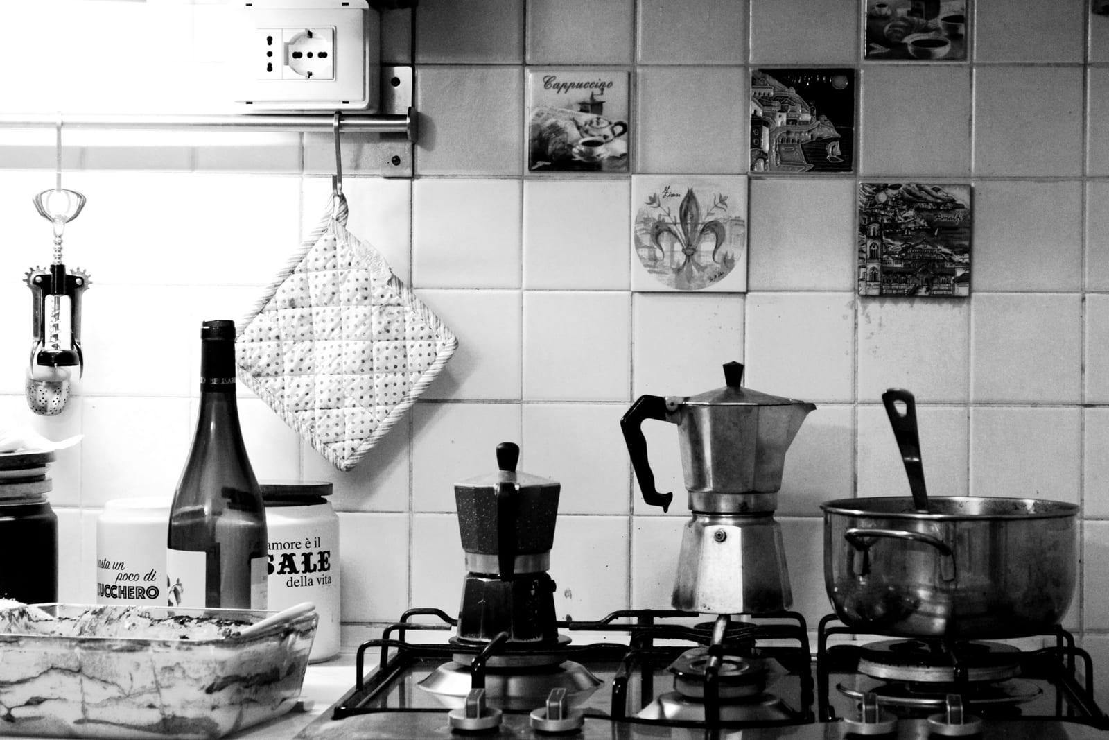 A home kitchen counter with two stovetop moka pots on a gas hob, beside jars and a tiled wall