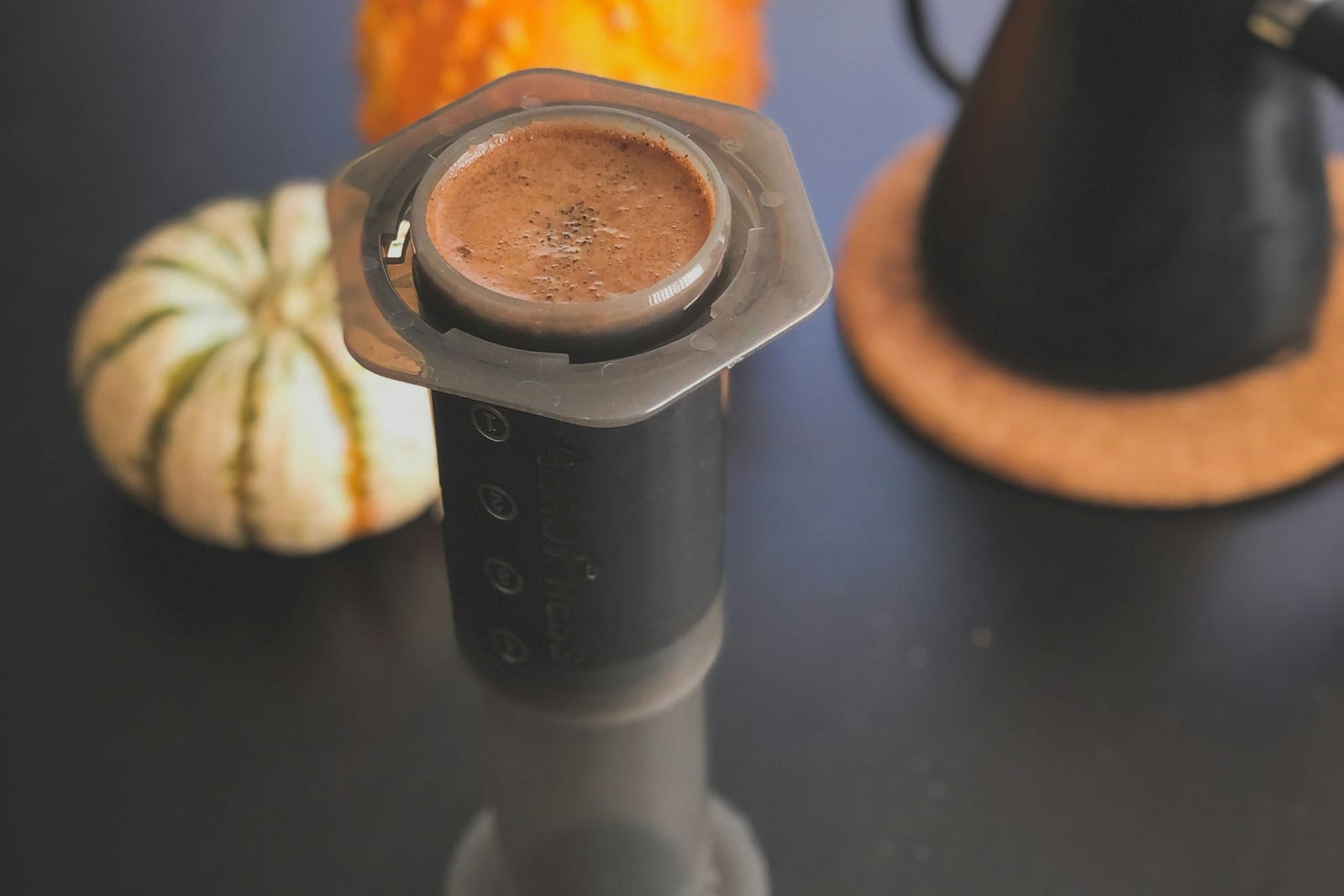 Un AeroPress invertido con el café molido medio-fino ya extraído, visible a través de la tapa del filtro, una calabaza ornamental rayada a la izquierda y una tetera negra de cuello de cisne a la derecha, sobre una superficie oscura