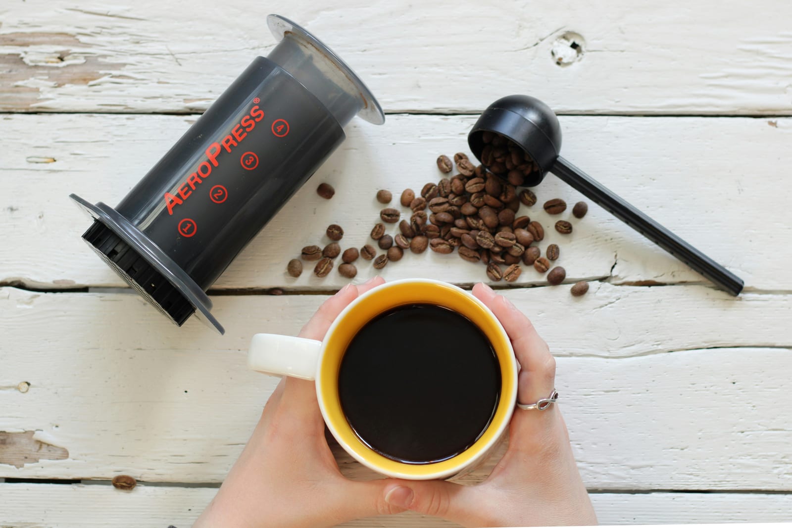 Manos sosteniendo una taza de café AeroPress recién preparado junto a una cucharada de granos de café con molienda medio-fina