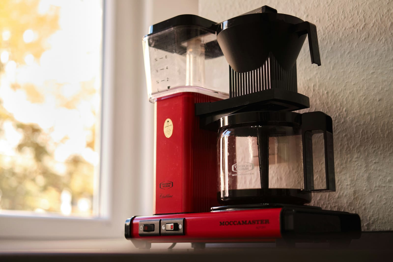 Uma cafeteira de gotejamento Technivorm Moccamaster vermelha ao lado de uma janela ensolarada, com café escuro na jarra de vidro embaixo do cesto