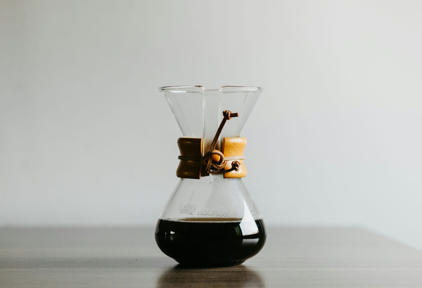 沙漏造型的 Chemex 玻璃壶,带木质环和皮绳,冲好的咖啡盛在壶身,摆放在木质桌面上,背景是一面柔和的灰白色墙面