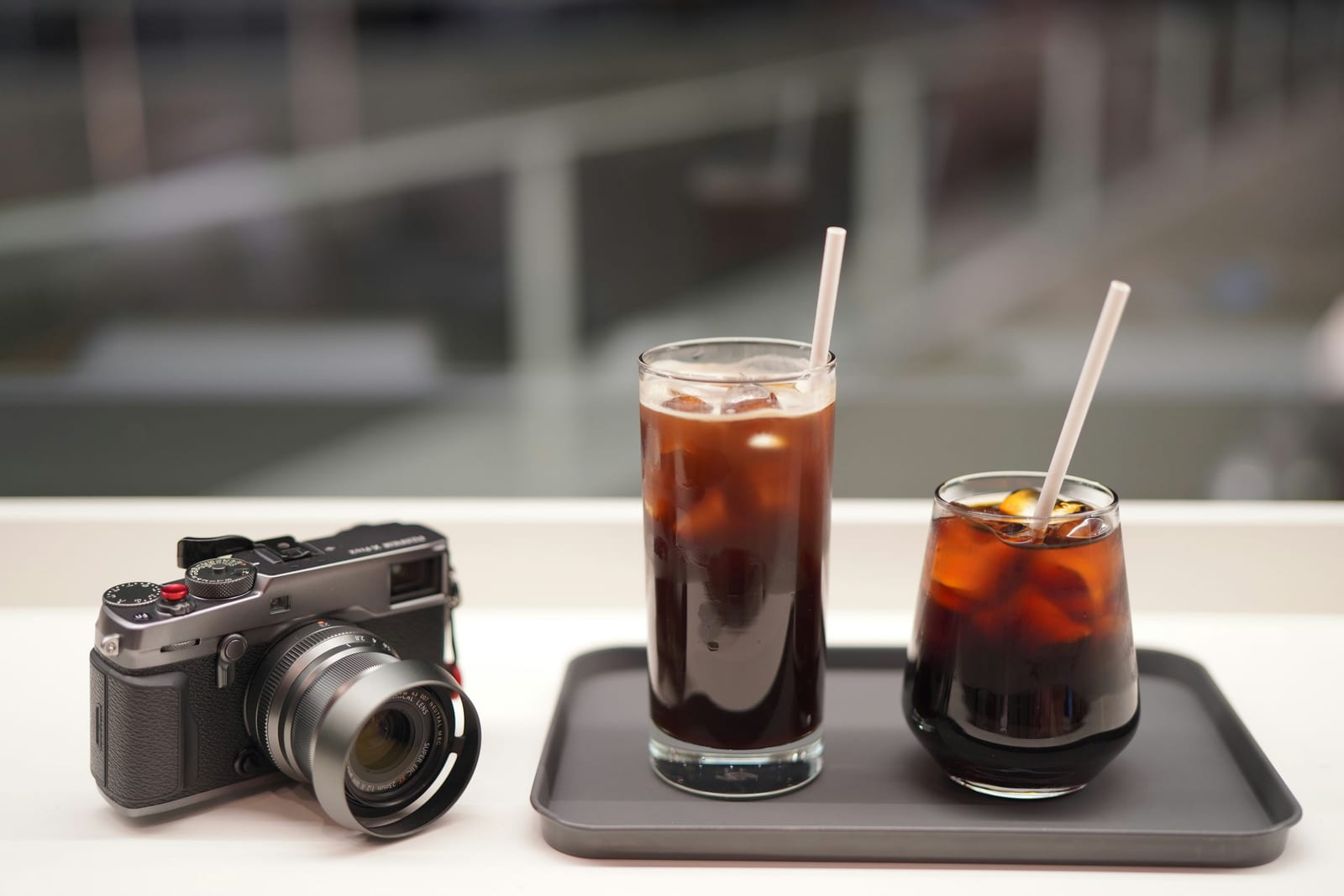 Dos vasos de cold brew helado sobre una bandeja gris al lado de una cámara fotográfica telemétrica Fujifilm vintage, el concentrado de molienda gruesa diluido sobre hielo