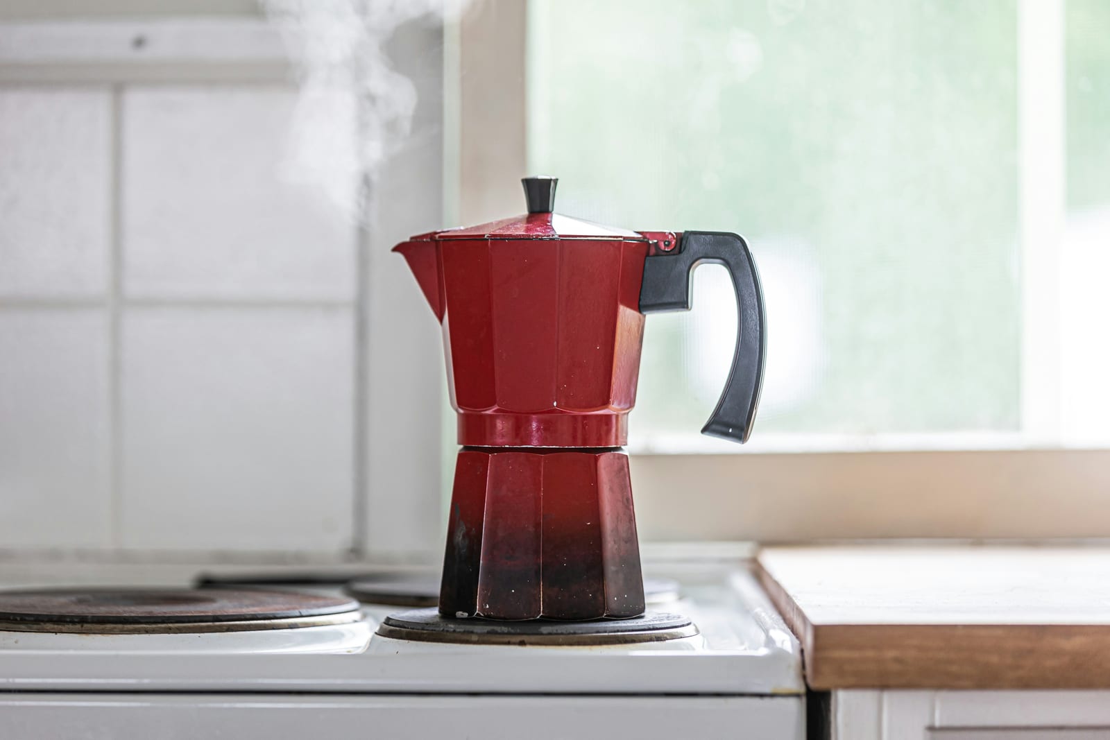 Una cafetera moka roja sobre la estufa con vapor saliendo del pico, la señal de que el café molido fino termina su extracción a presión