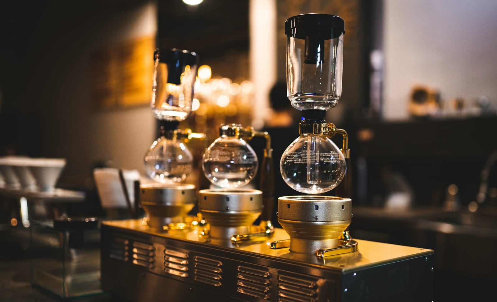Una hilera de tres cafeteras de sifón de cristal sobre una base de quemadores de metal pulido en una cafetería, iluminación cálida de fondo