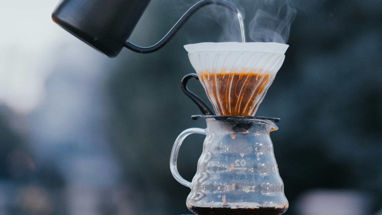 A hand pouring water from a black gooseneck kettle into a paper-filtered V60 cone over a glass carafe, steam rising, medium-fine grounds blooming